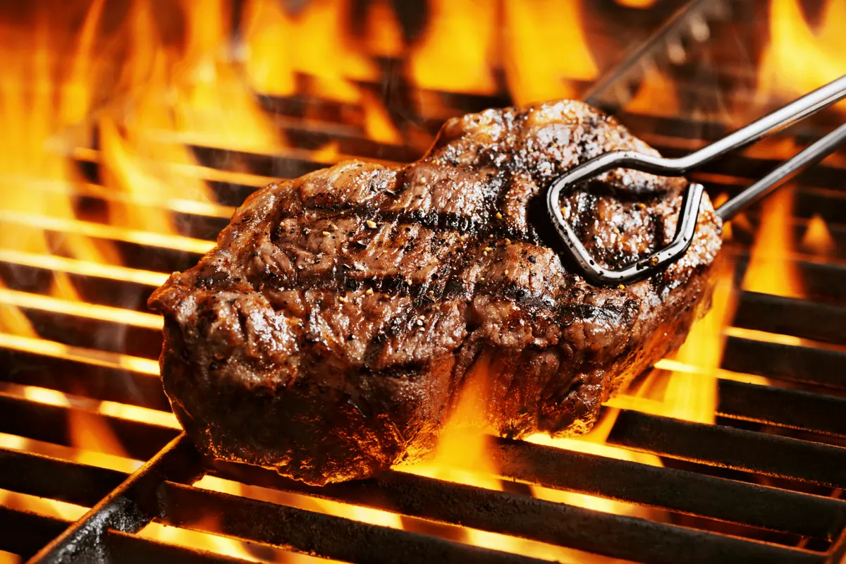 A steak turned with tongs over open flames on a barbecue