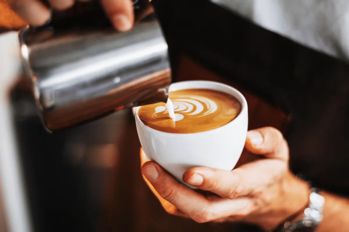 A barista’s hands pouring milk into a flat white, latte art forming in the cup