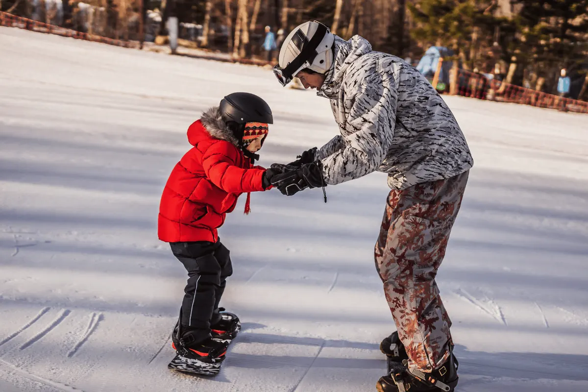 A father guiding his child down a slope on a snowboard