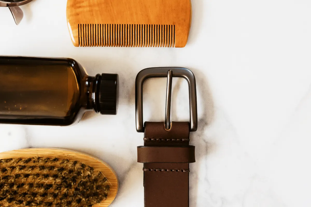 A comb, bristle brush, leather belt and amber bottle laid out on marble
