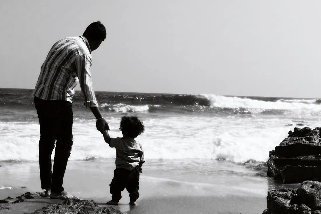 A father and small child walking hand in hand along the shoreline, in black and white