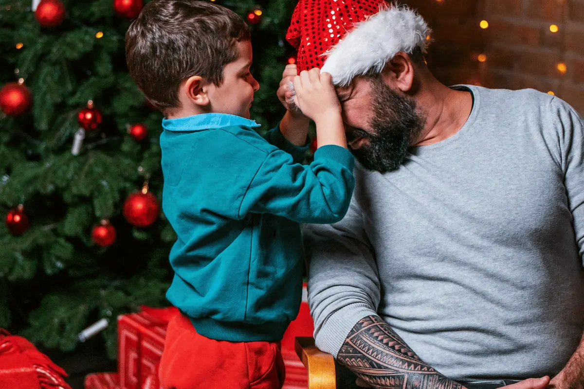 A father and son in Santa hats at the Christmas tree with presents underneath