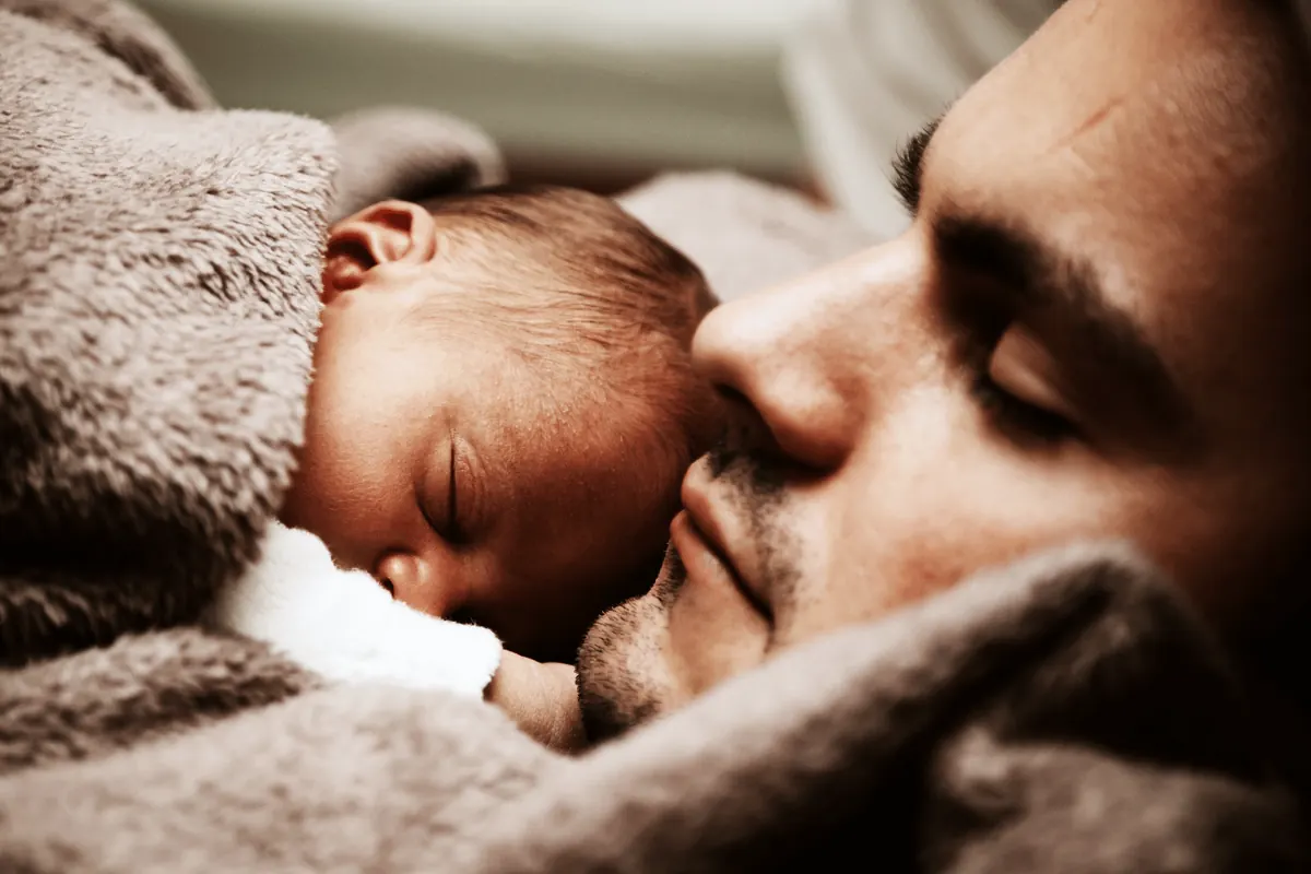A father asleep face to face with his newborn under a blanket