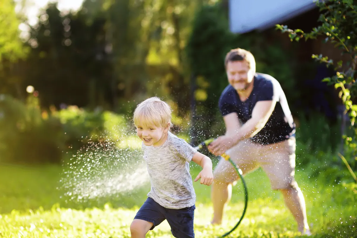 A father chasing his son with a garden hose across a sunlit back lawn