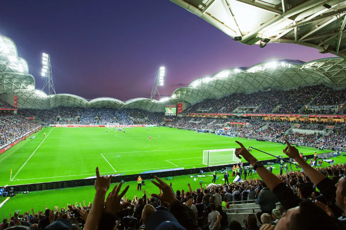 Supporters with their arms raised in a packed stadium at dusk, the floodlit pitch below them