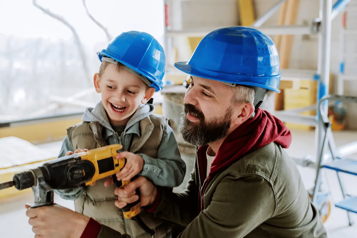 A father and son in hard hats using a cordless drill on a build